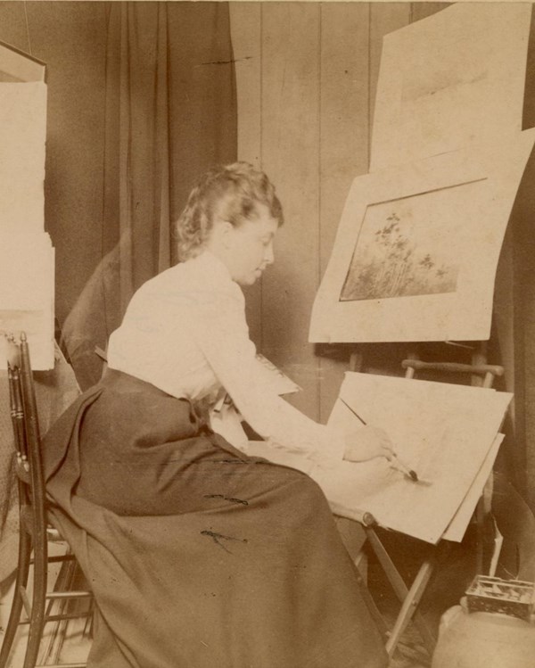 woman in white blouse and long dark skirt sits painting at easel surrounded by art