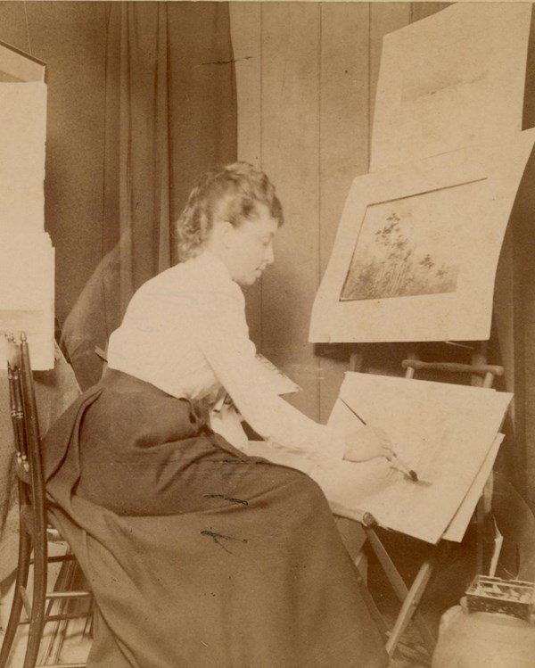 woman in white blouse and long dark skirt sits painting at easel surrounded by art