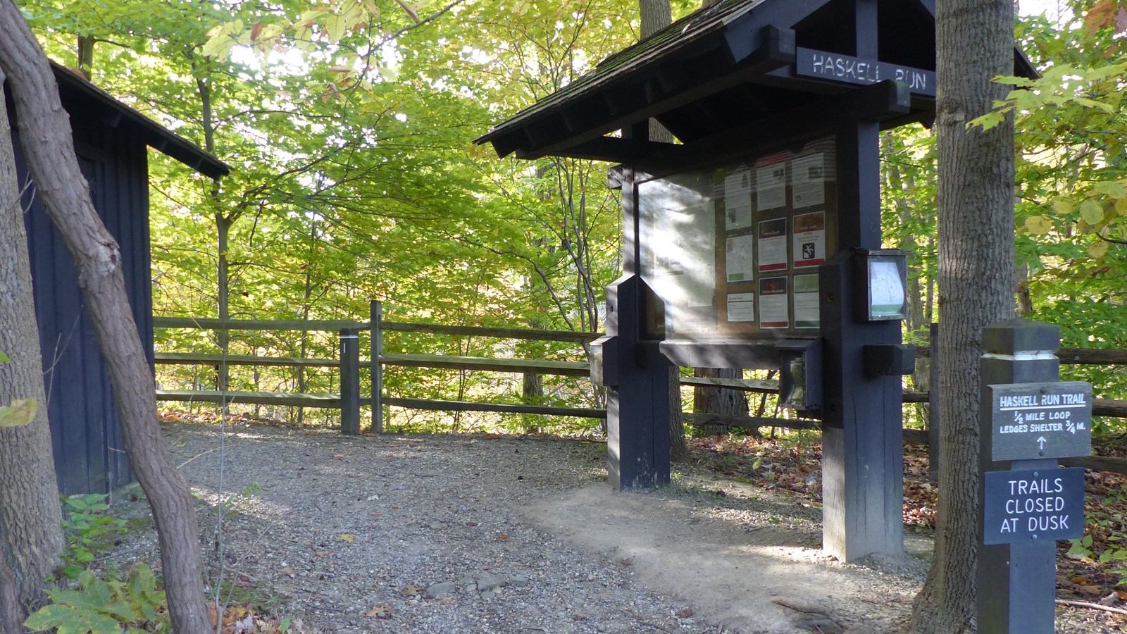 Unpaved trail starts between “Haskell Run” bulletin board and a shed, and bends left along a fence.