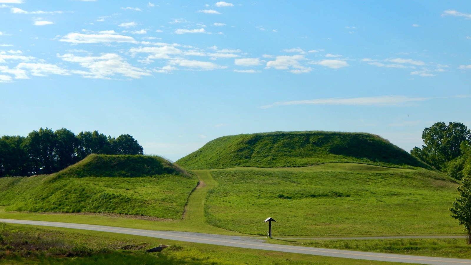 Two grassy mounds in an open field with the park road running in front of them.