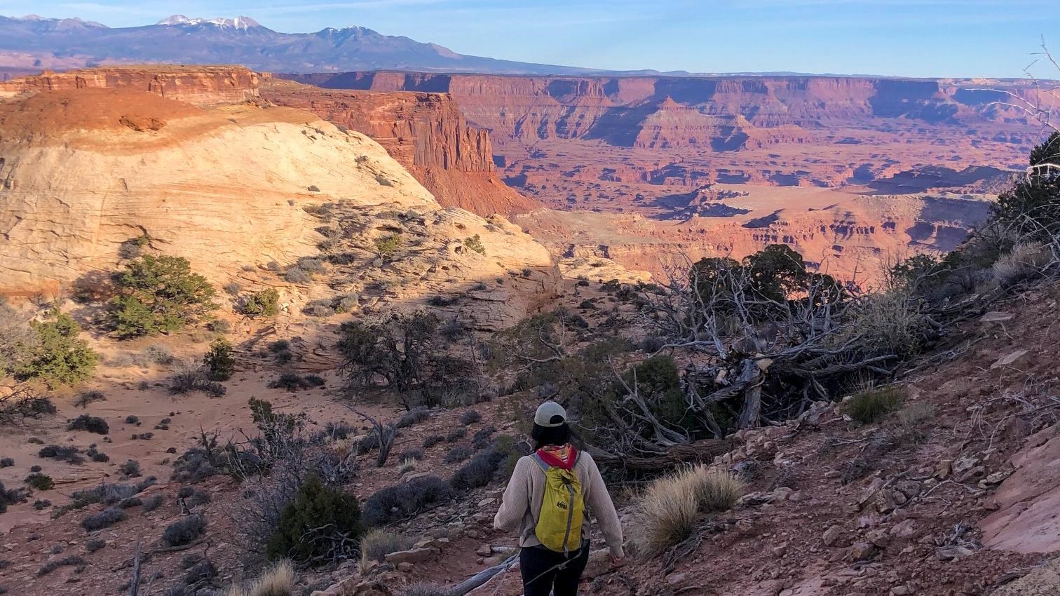 A hiker descends the trail into a canyon, sweeping views are visible in the distance. 