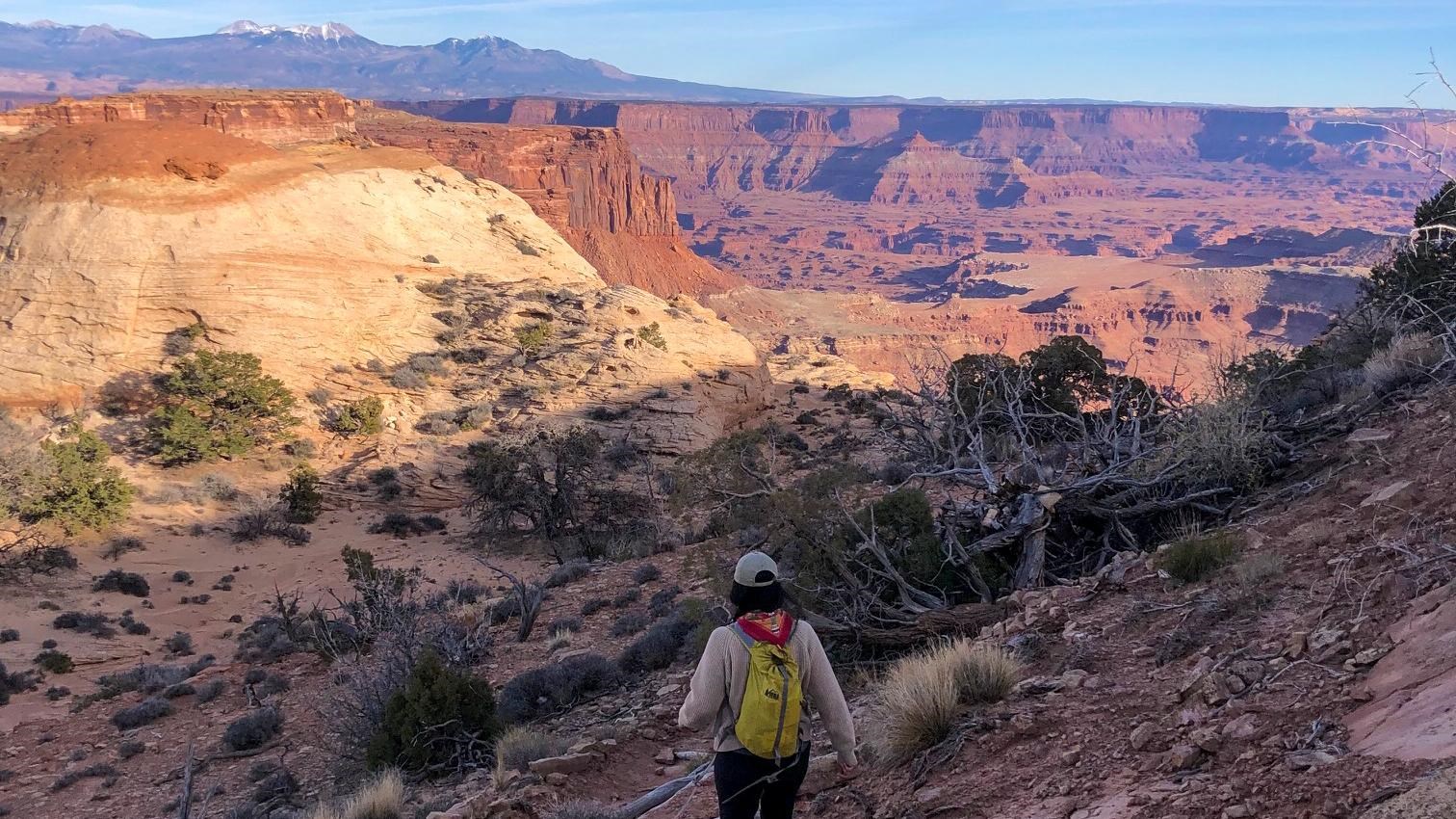 A hiker descends the trail into a canyon, sweeping views are visible in the distance.