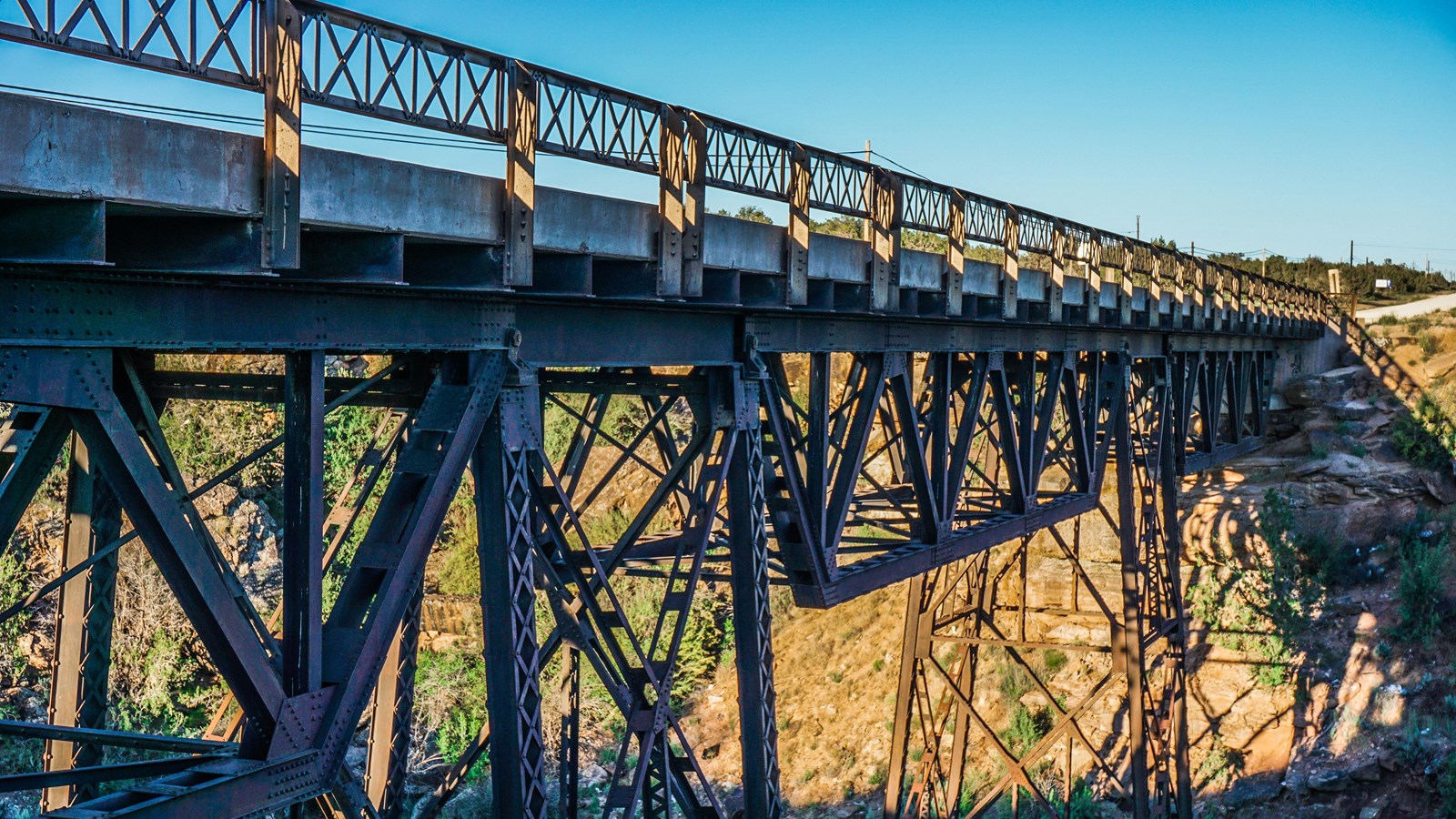 A large steel truss style bridge with red rocks, green shrubs, and blue sky behind.