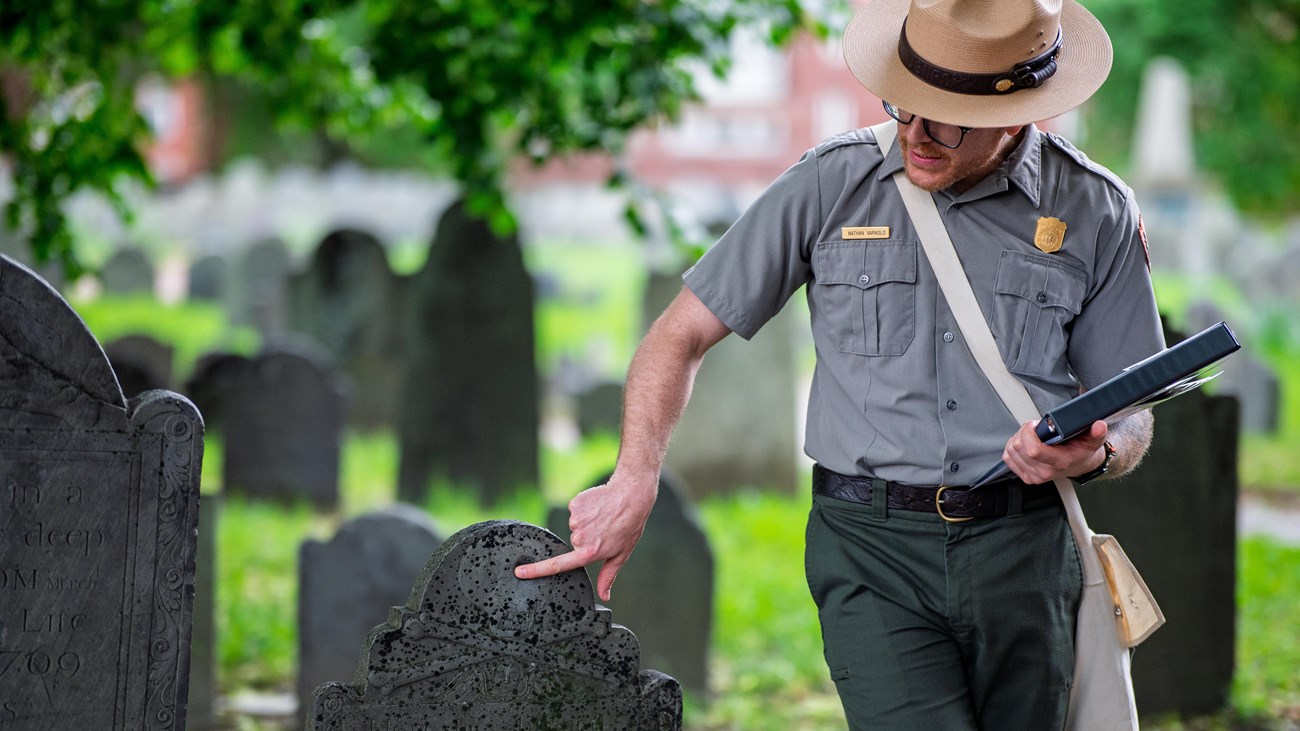 A park ranger points out something on a historic 18th century gravestone