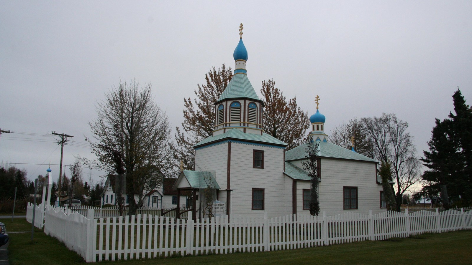 A small white Russian Orthodox church with a blue dome and surrounded by a white picket fence.
