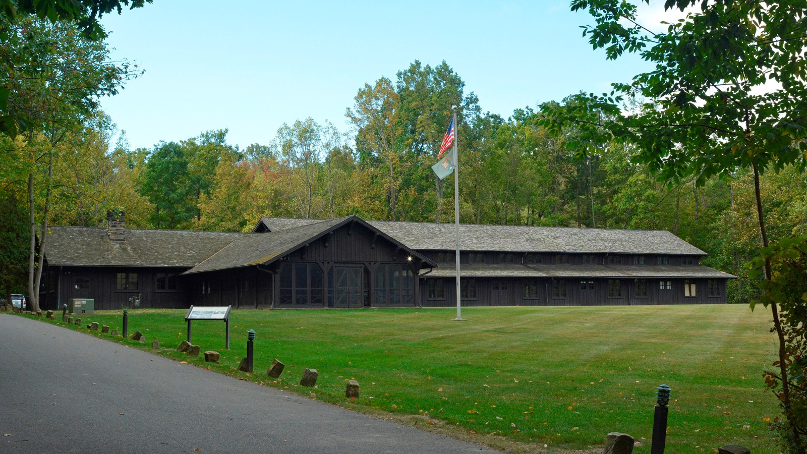 Long, brown, rustic building with porch overlooks play field with panel by a driveway and flagpole.