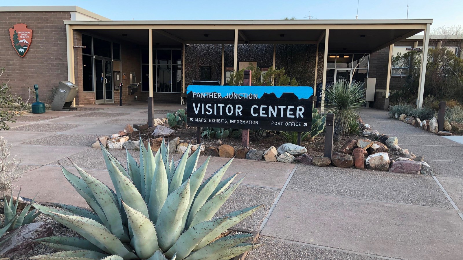 A large agave is growing in front of a square building with a NPS arrowhead on the side of the wall.