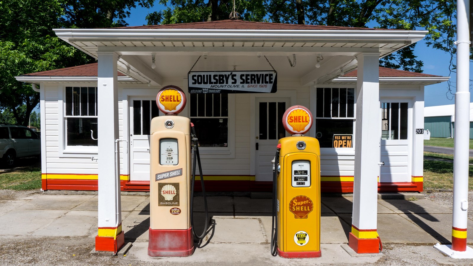 A white building with a tan roof with two old-fashioned yellow and tan shell gas pumps out front.