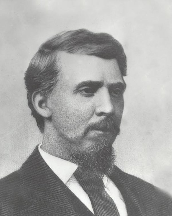 White man with mustache and goatee wearing a black suit and tie with a white shirt.