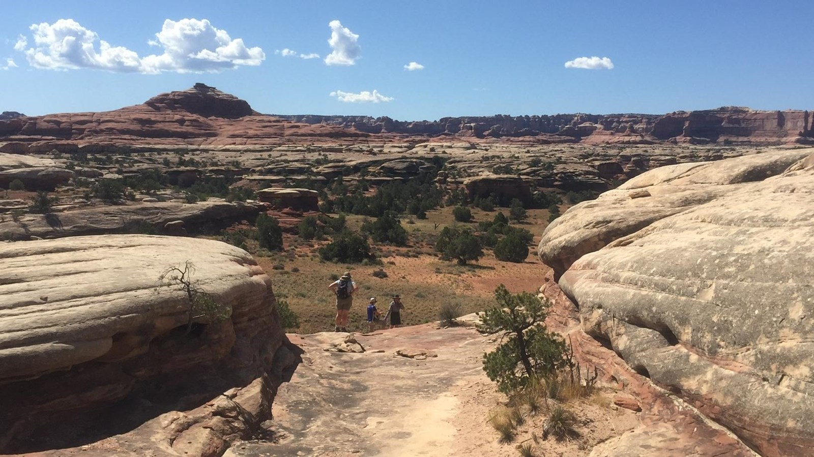 Two hikers are seen in the distance before descending into a canyon on a bright, bluebird day