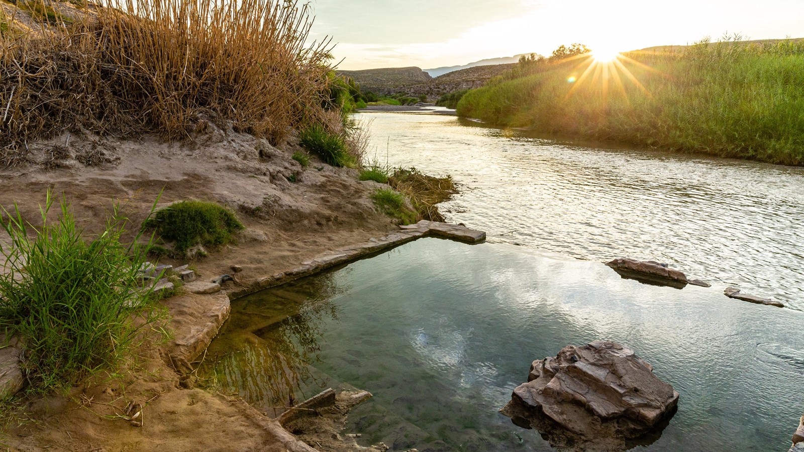 A pool of water encircled by a square rock foundation rises above a river.