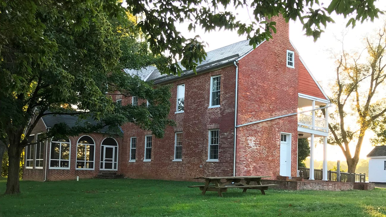 A picnic table sits on a green lawn with a large,P red brick house behind it.