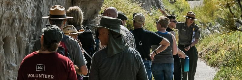 A park ranger points to a cliff face while a crowd of people gathers around. 