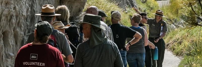 A park ranger points to a cliff face while a crowd of people gathers around.