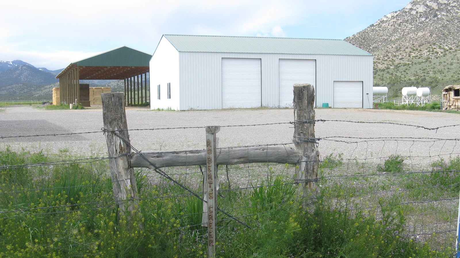 A silver-colored post stands in front of a wire fence with ranch buildings in the background.