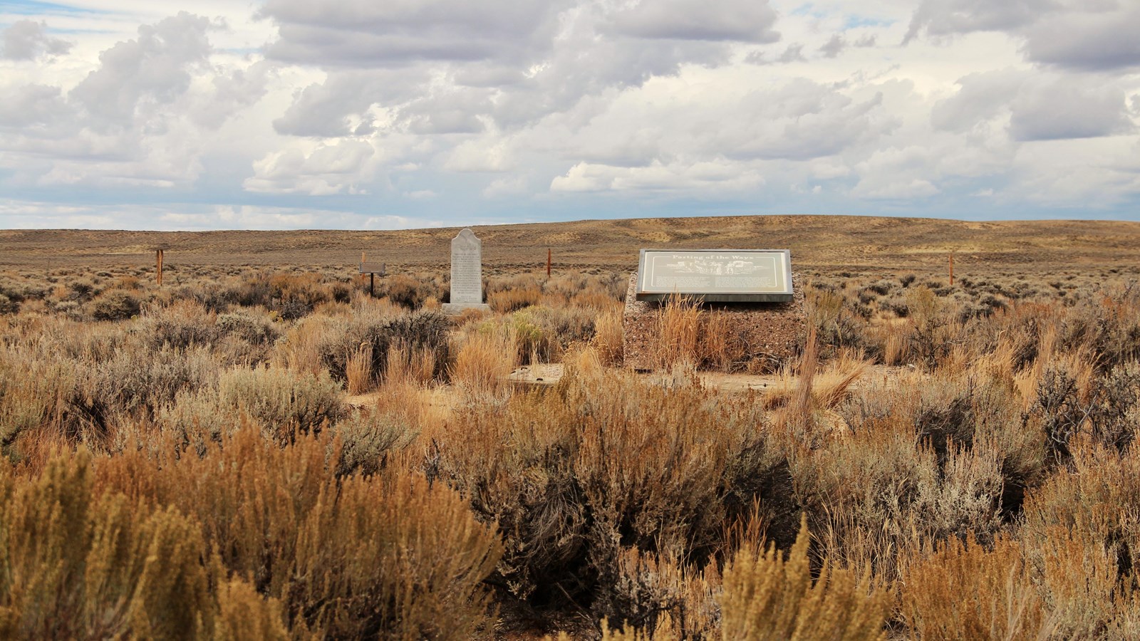 Sagebrush, wayside exhibit, granite monument, sign, fence, plain, and cloudy sky are visible