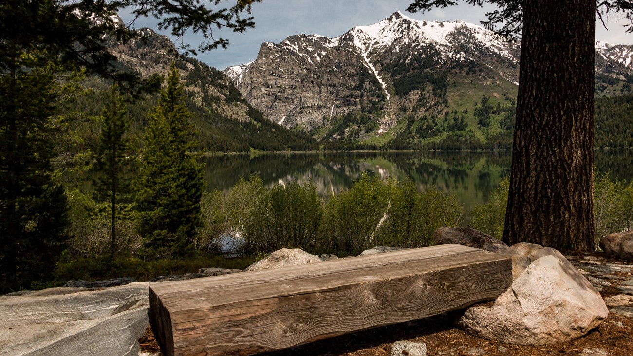 A wooden bench sits beside a lake surrounded by mountains.