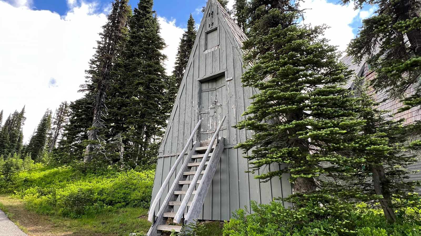 A-frame building with steep roof, wooden paneling, stairs leading up to door, surrounded by trees