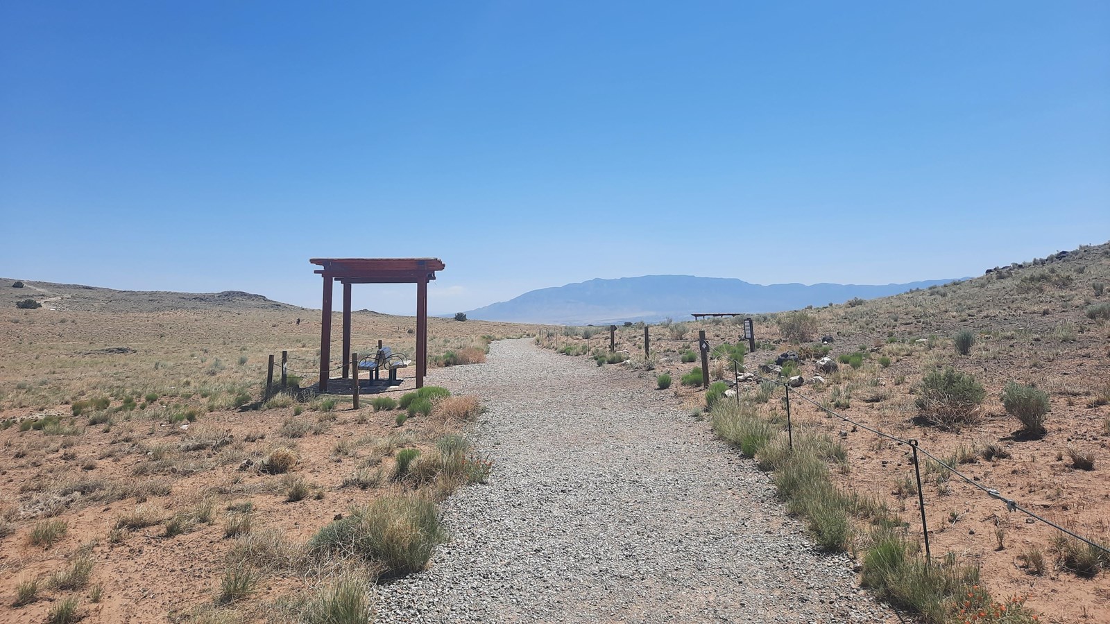 A trail through a desert landscape with a shade structure and a bench next to it.