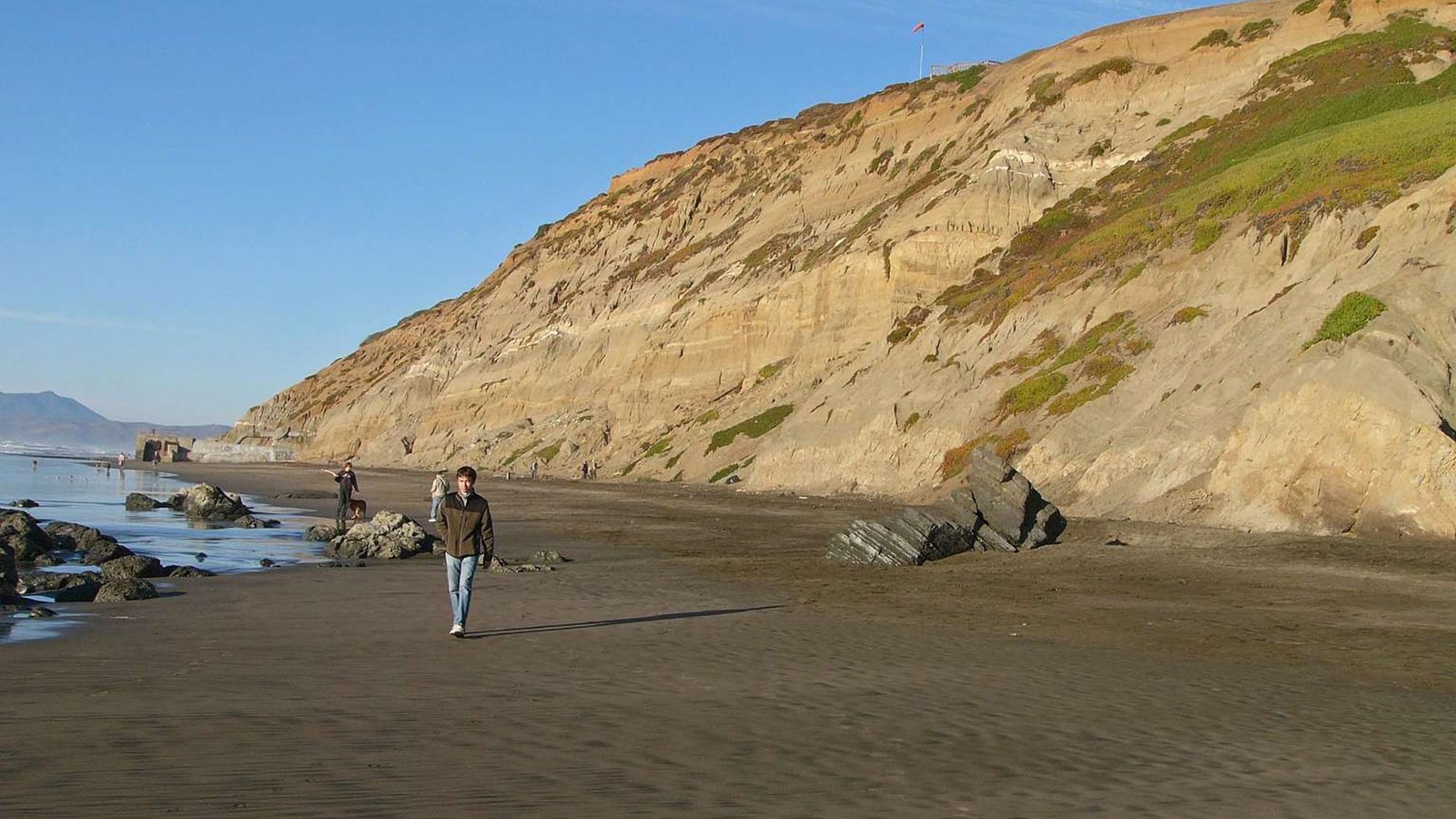 The tall, tan cliffs of Fort Funston with people walking along the seashore near their base.