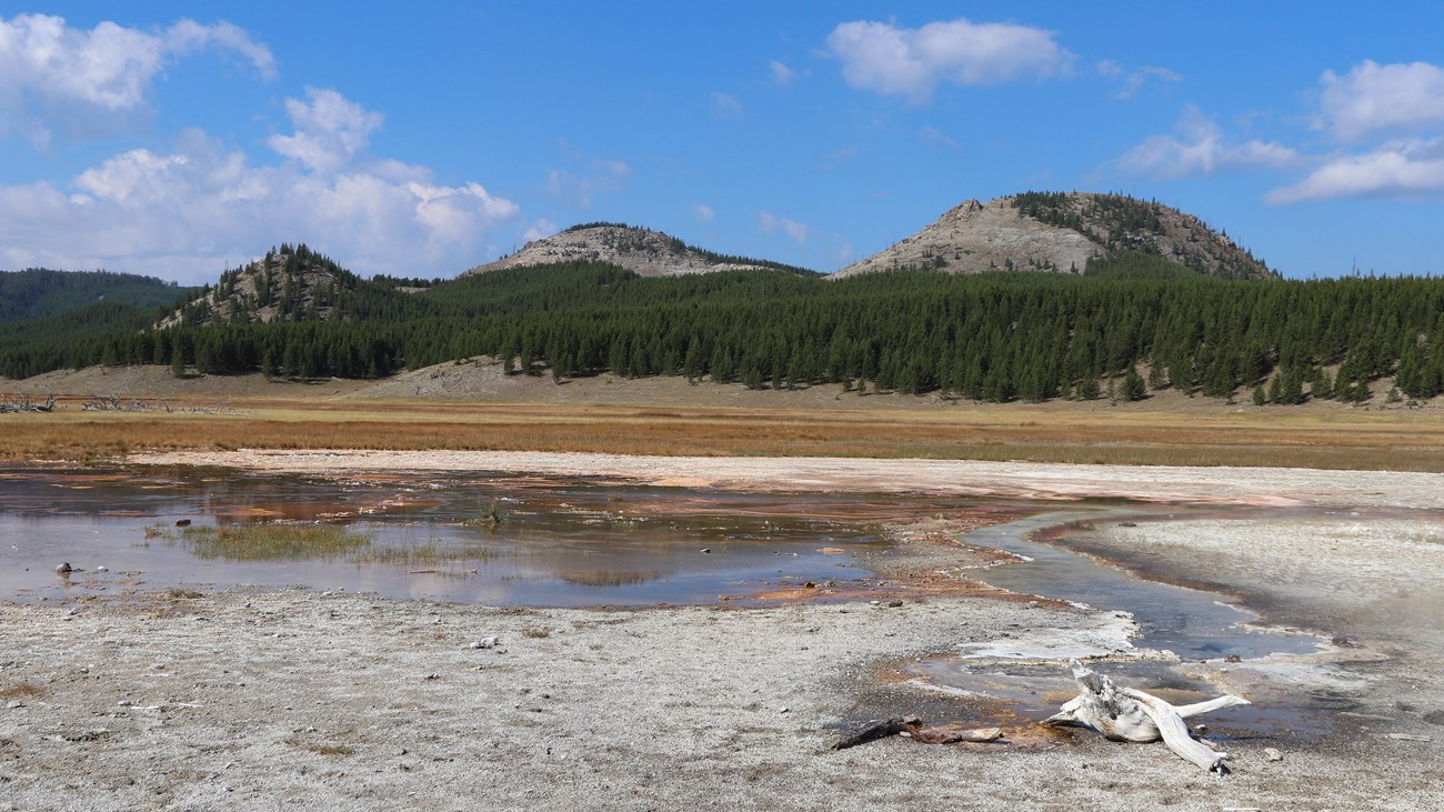 A thermal area sits in a meadow in front of rolling hills.