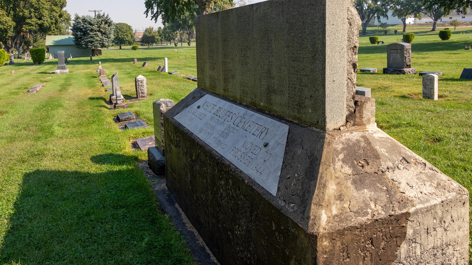 Color photograph of a row of graves in a green cemetery. In the foreground is a large stone monument