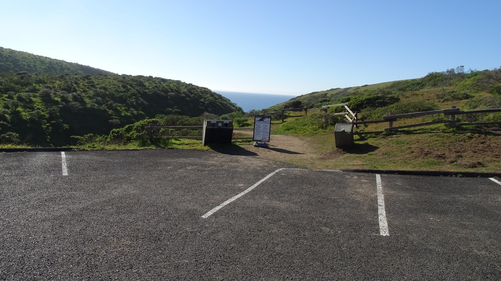 A dirt trails departs from a paved parking lot toward the ocean in the distance.