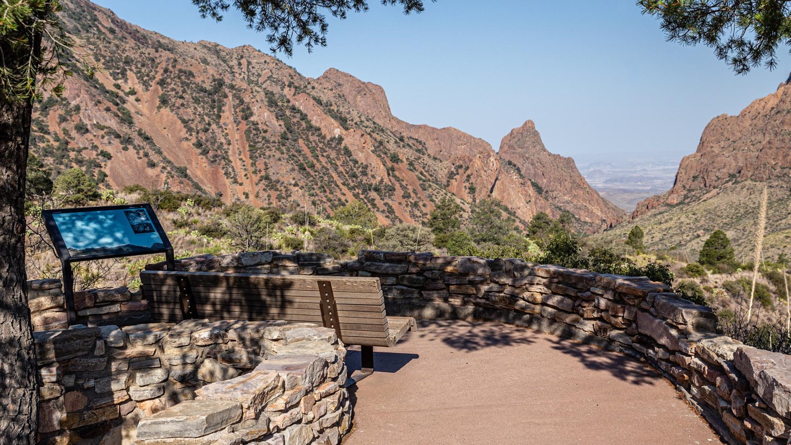 A sign with a metal frame is perched on a low rock wall looking out on the Chisos Mountains.