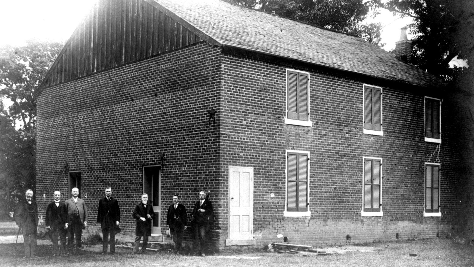 Six men in suits stand in front of a two story brick church, historical photo.