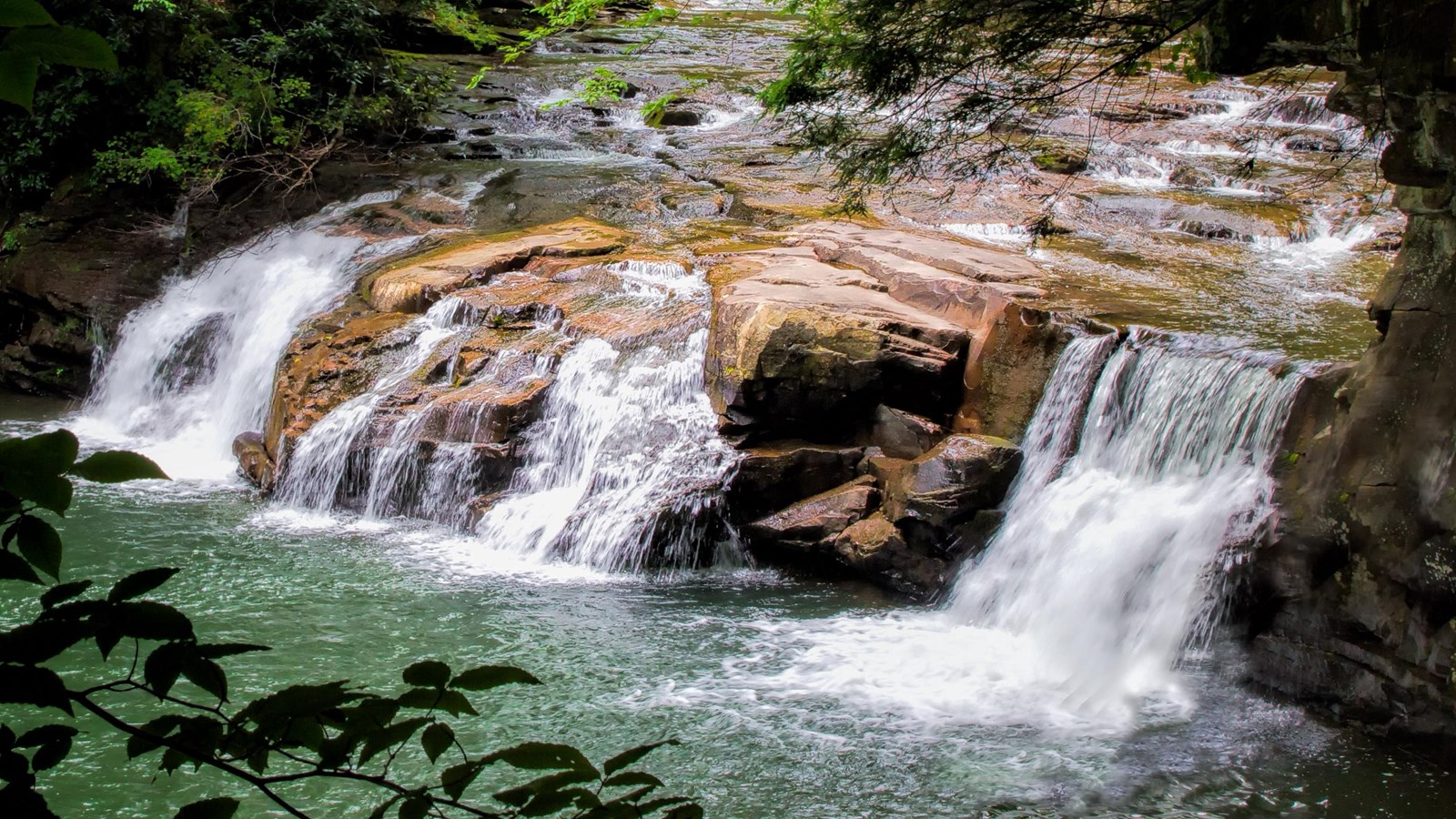 Water cascading over wide reddish-brown rocks into a deep pool.