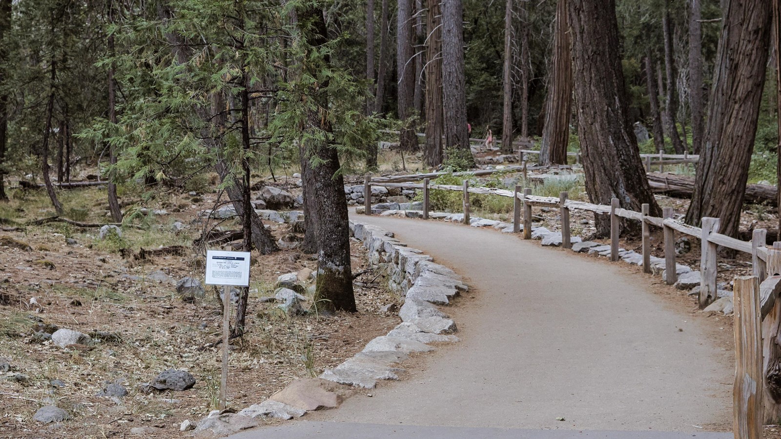 A paved path leads through a treed area lined with wood barrier fencing and boulders