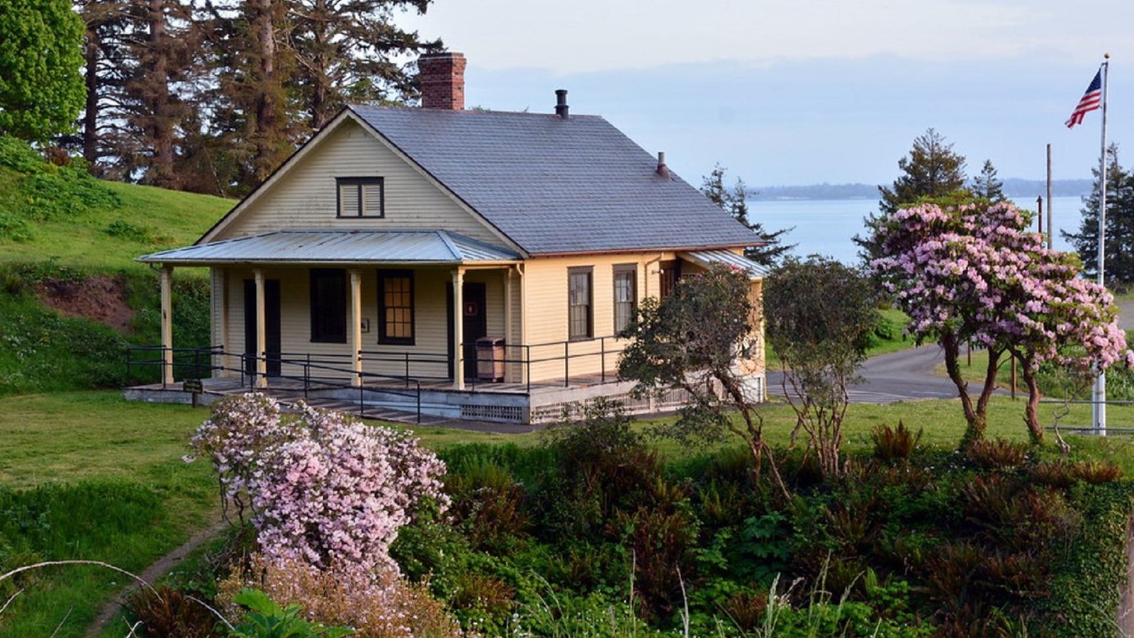 A small tan building with a covered porch surrounded by grass and flowering trees, ocean behind