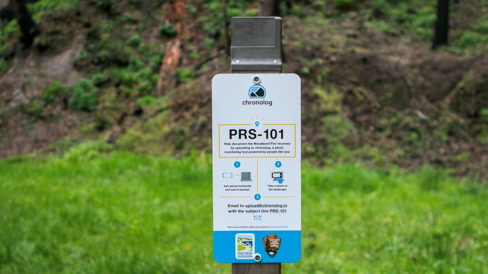 A white and blue sign with grey photo bracket in front of green grass and a forested hillside.