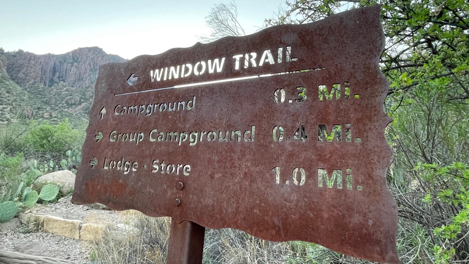 A metal sign has the names of trails and their distances cut into it.