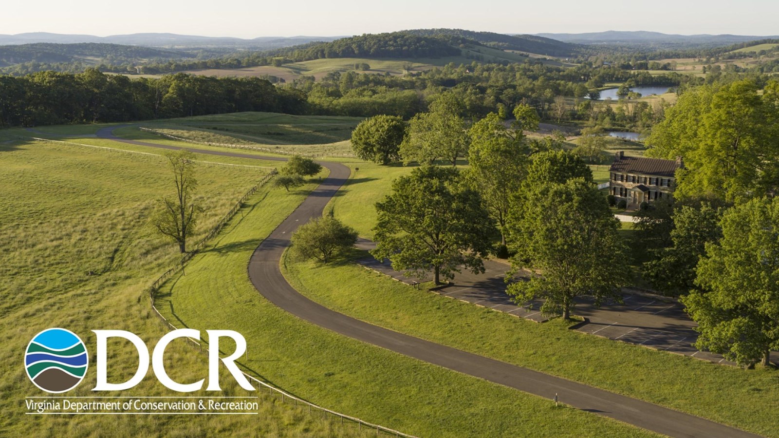 A road passes through a green landscape with mansion on the right, and a river in the background