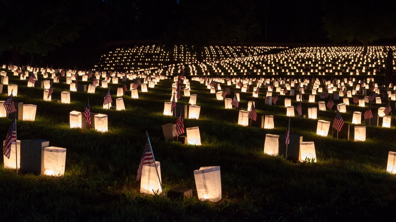 Individual luminaries sit in front of hundreds of graves in the rolling hills of a cemetery.