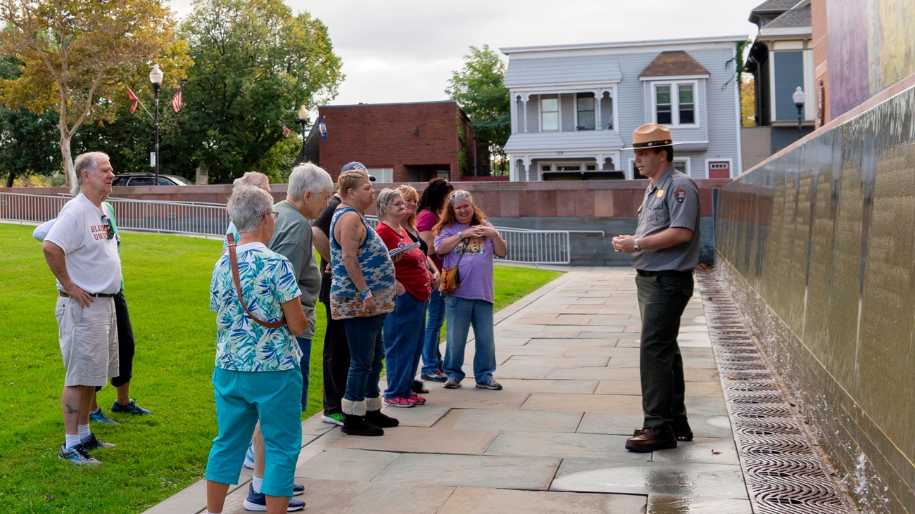A ranger stands and speaks to a group of people in front of a wall.