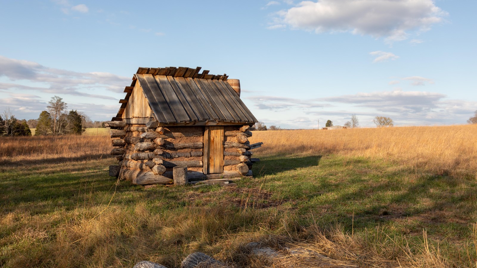 A wood log house sits in the middle of a grassy field. Logs sit on the ground in the foreground. 
