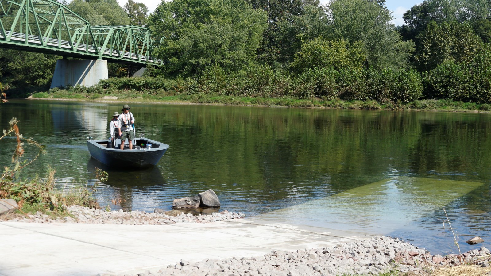 View of concrete ramp leading into the river from shore. A boat approaches ramp. Bridge behind them.