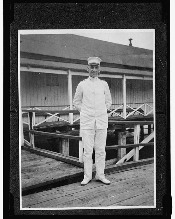 A man stands in a white uniform and hat with his hands behind his back.