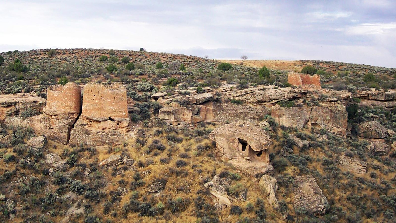 Remains of a stone structure tucked underneath a large boulder. The boulder sits on the side of a ca