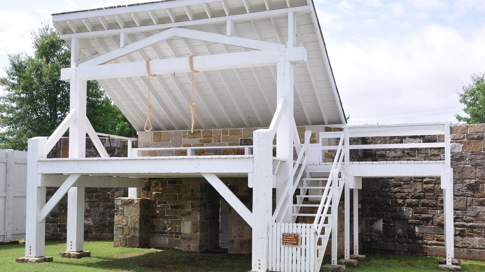 Two nooses hang from the crossbeam of the white gallows structure. Surrounded by a fence. 