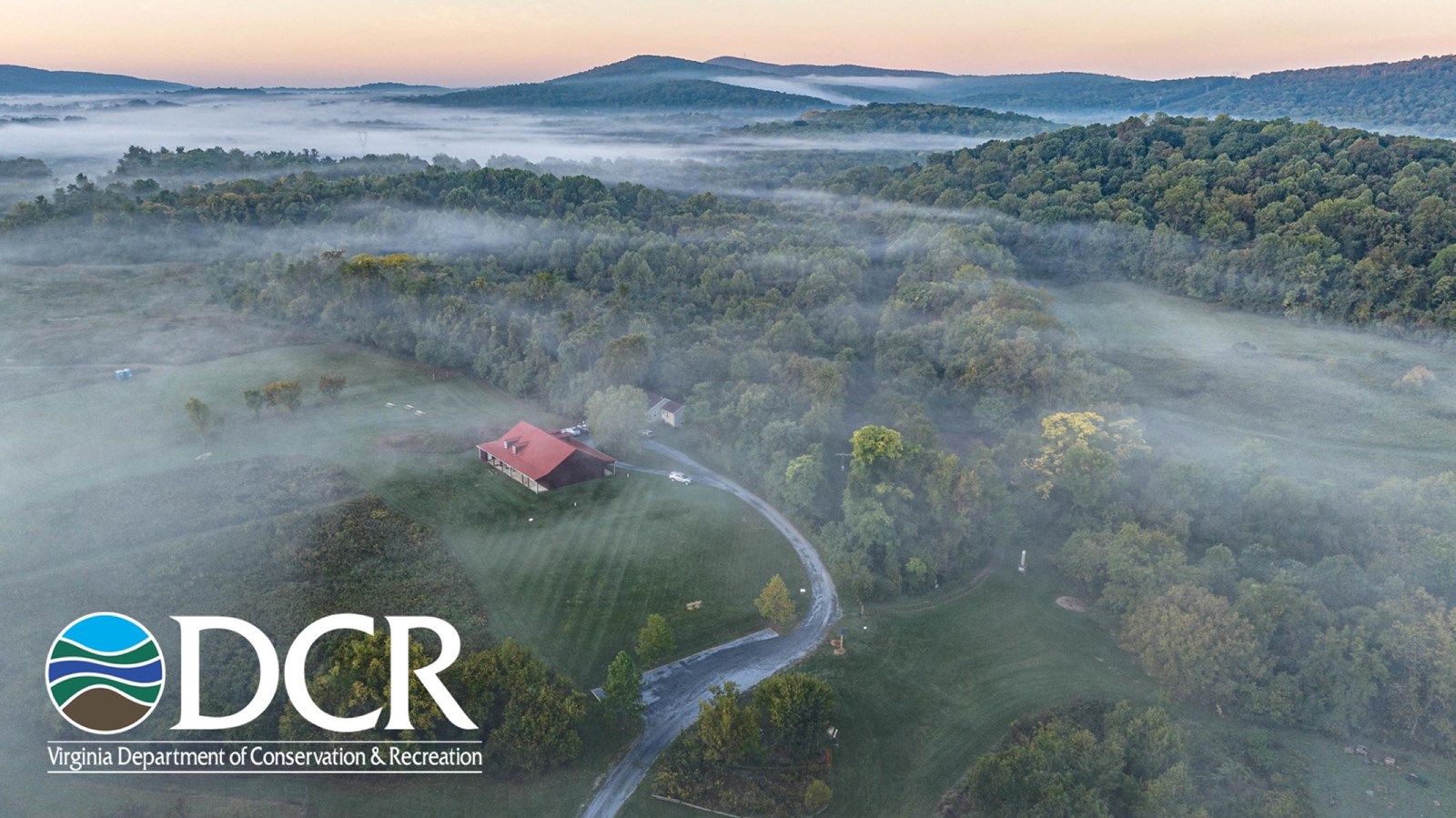 A misty landscape at dawn with a red-roofed building and road surrounded by trees and hills.