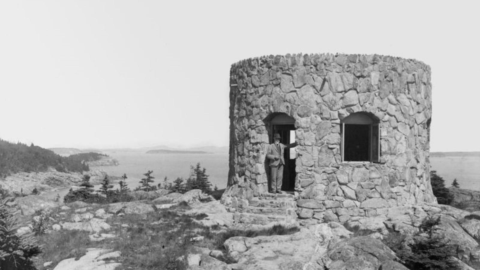 A person stands in a cylindrical stone house overlooking a coastal view.