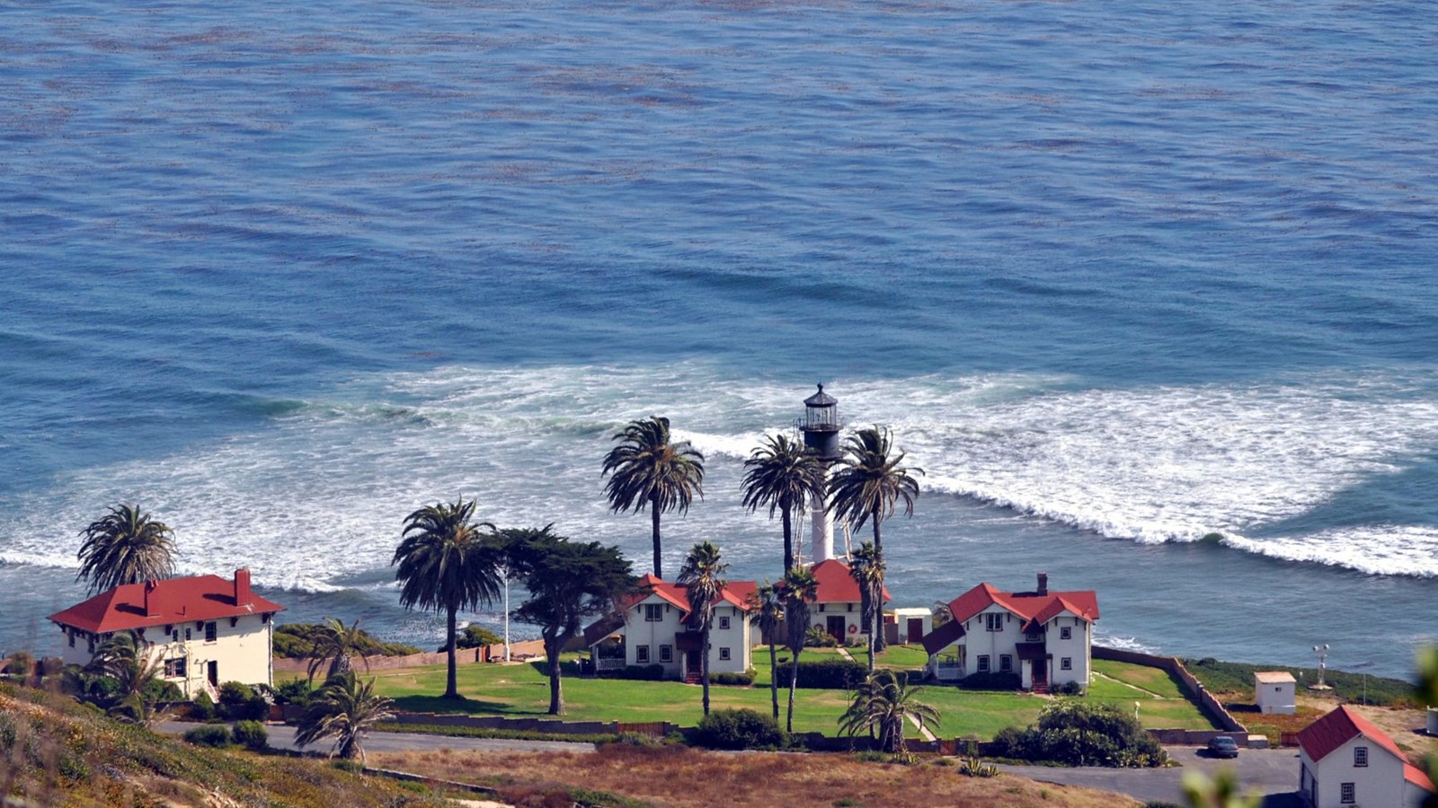 Color photograph of a lighthouse tower surrounded by white buildings with red roofs and the sea