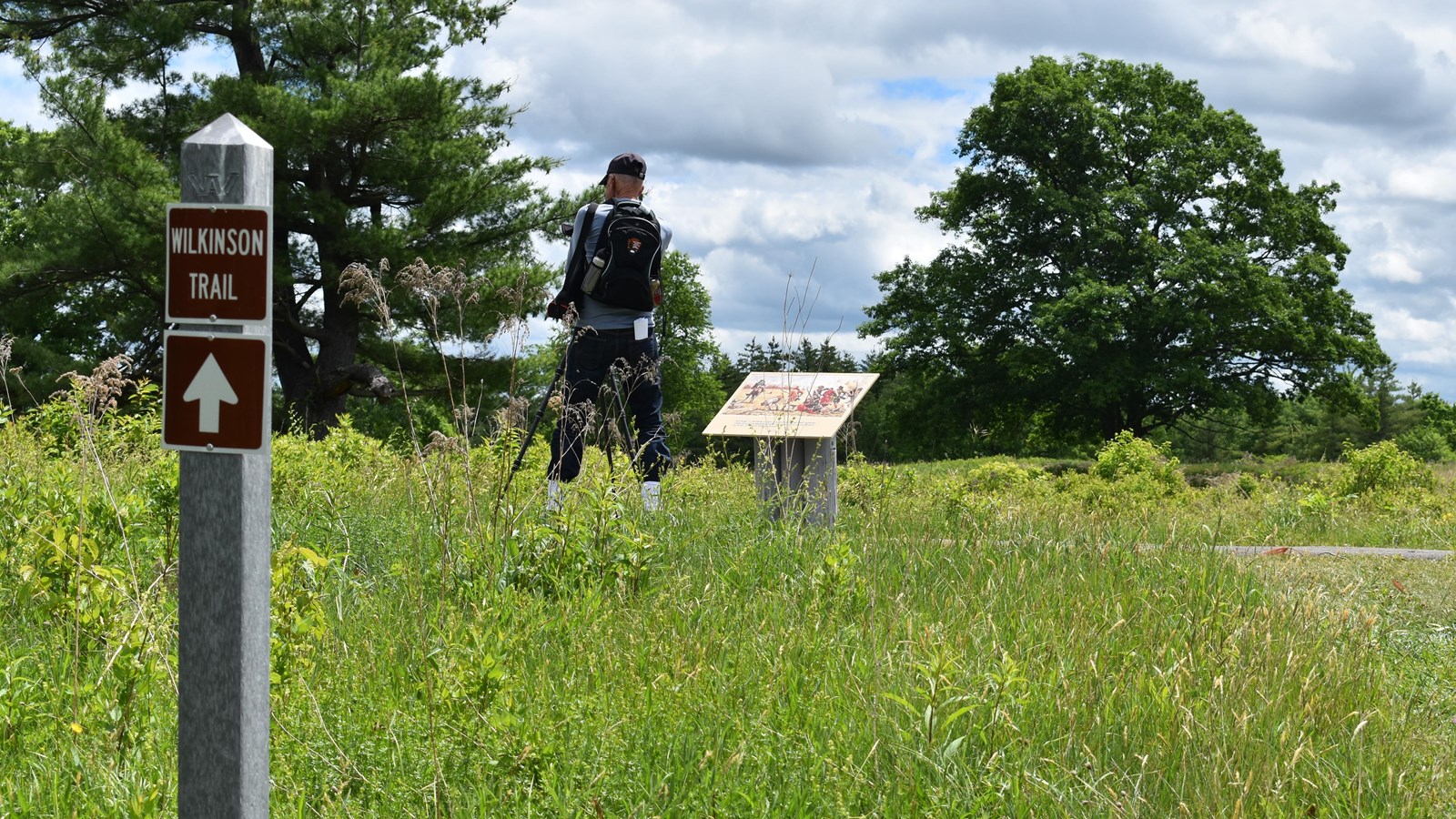A gray post marker for the Wilkinson Trail. In the near distance, stands a man in front of a wayside