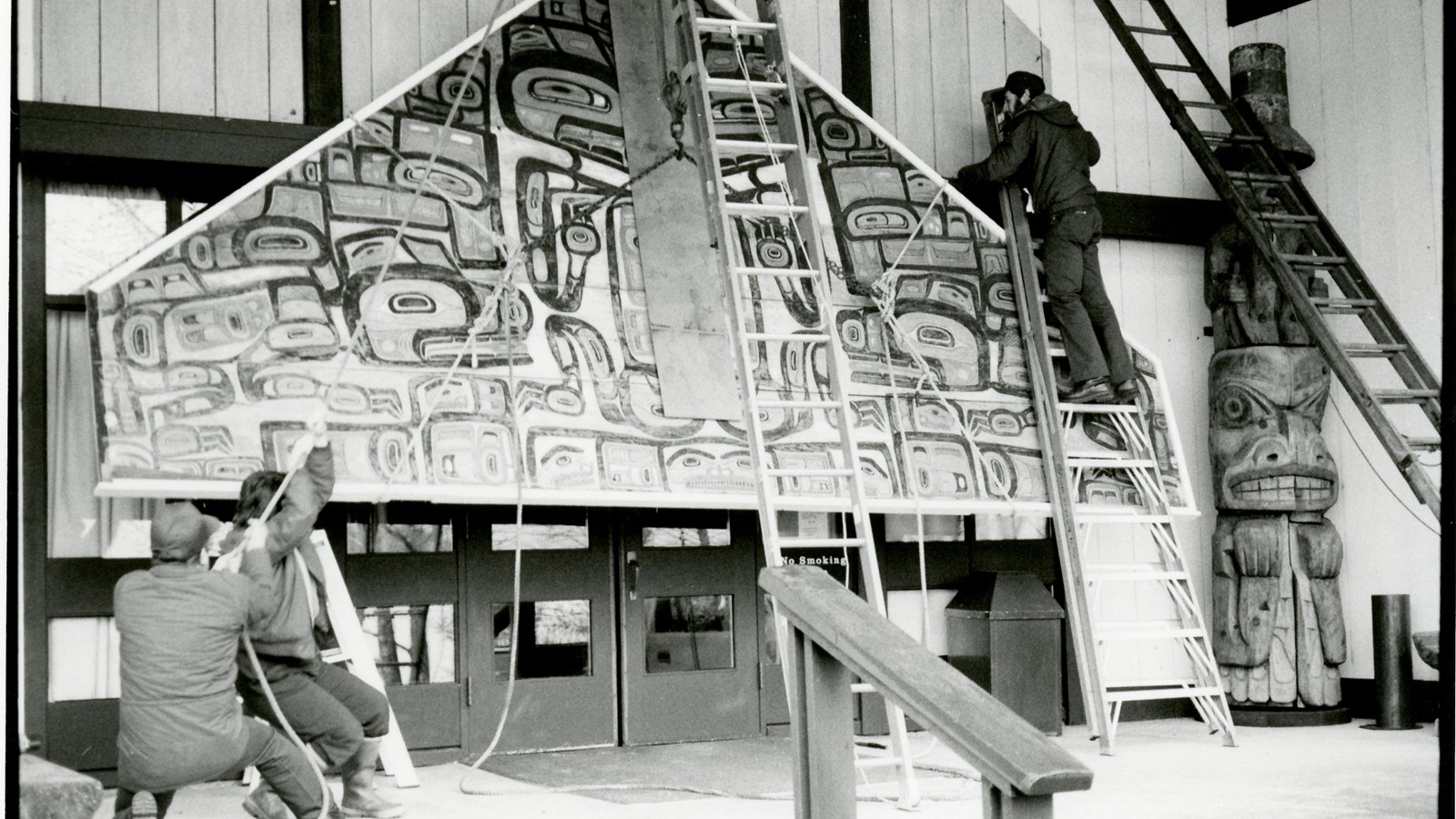 A black and white photo of workers with ropes and ladders lowering a large outdoor panel.