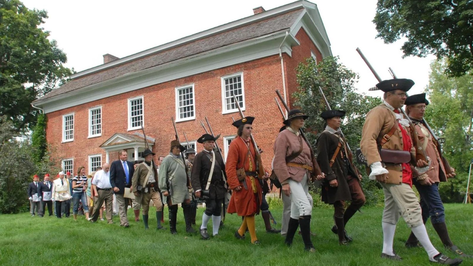 A line of people dressed as 18th C militia and in modern clothes march past a large brick house.