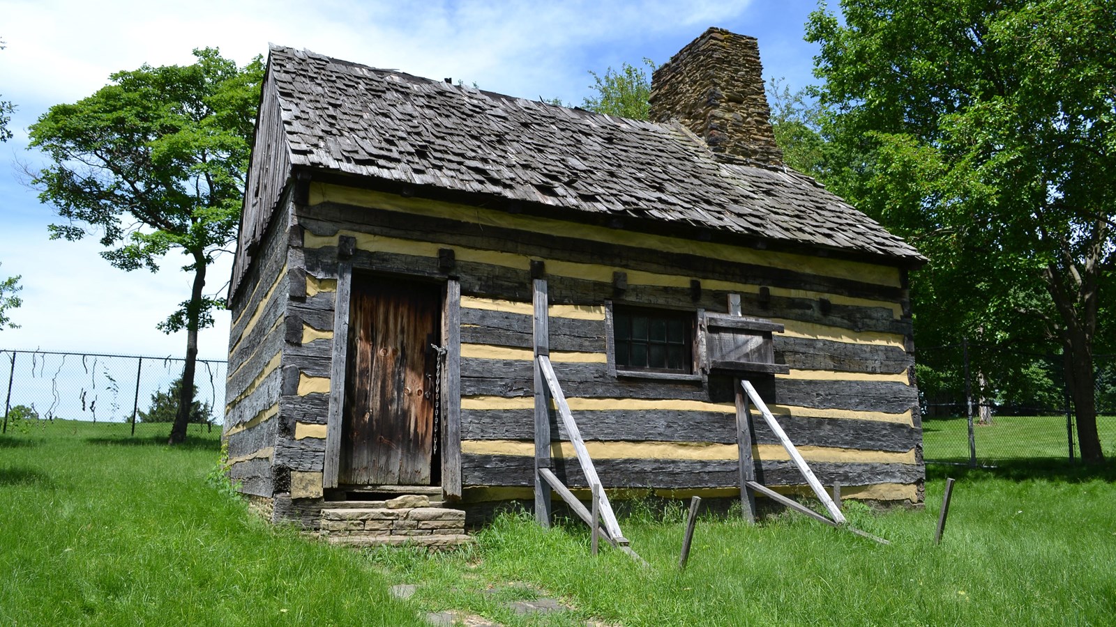An 18th century log cabin in a grassy field with a fence behind it