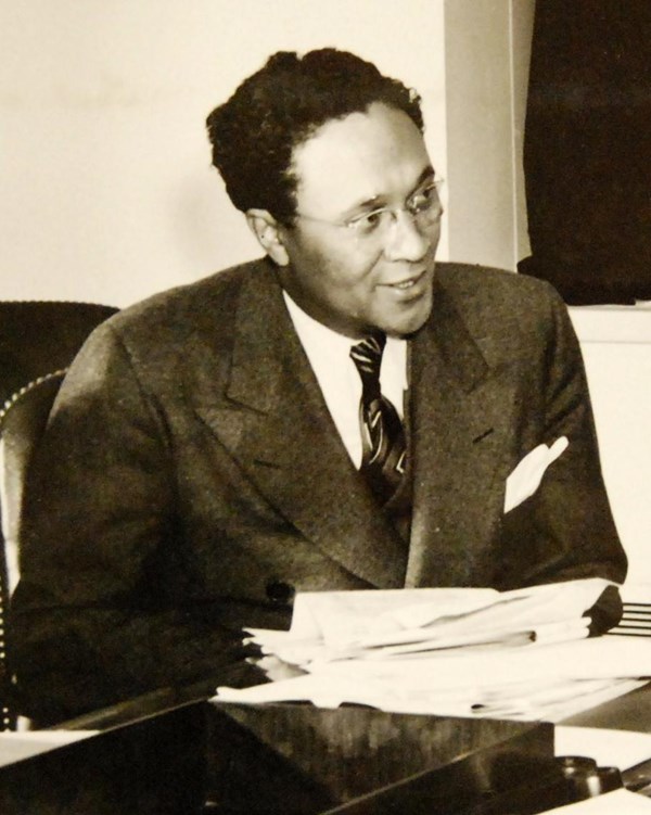 Black man wearing suit, tie, and glasses seated behind messy stack of papers